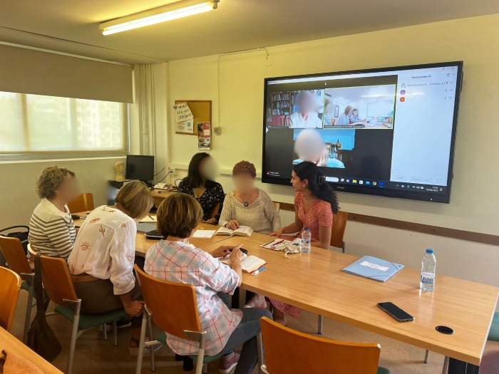 Six people, including Alana, are gathered at a conference table with a large mounted TV in the background showing a couple of Zoom participants in a virtual meeting room. Faces other than Alana's are blurred for anonymity.