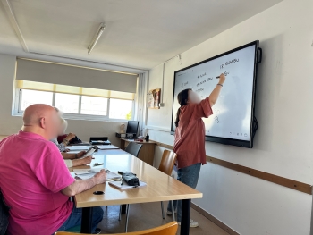 The photo shows up close a teacher at a white board writing something, face blurred and a few students in the front row listening, also with blurred faces.