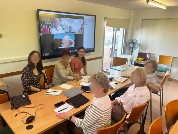 Six people, including Alana, are gathered at a table with a large mounted TV behind them showing two people participating virtually