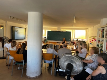 The photo shows a room of people seated at chairs around tables participating in a discussion. There is a large white fan near the back of the room, close to the camera's point-of-view.