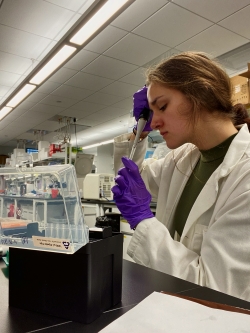 Alex works in a lab wearing purple plastic protective gloves. In her right hand positioned vertically is some kind of extraction tool, which she is focused on using.