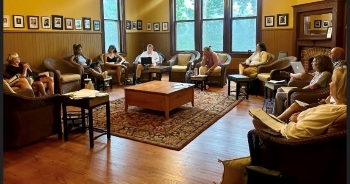 A formal, large study with enough room for dozens of people holds, instead, a broad circle of 10, all seated in arm chairs with laptops, tablets, or printed stacks of paper. All heads and eyes are turned toward Abi, who is seated on the far left and is reading.