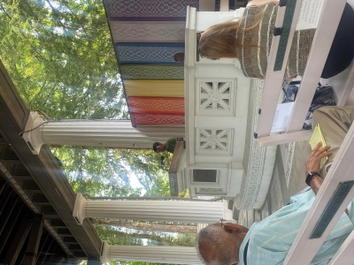 View from the audience near the front and stage left in an ampitheatre-style partially indoor, partially outdoor enclosure. Someone stands at the podium speaking, and there are rainbow-striped panels behind them.