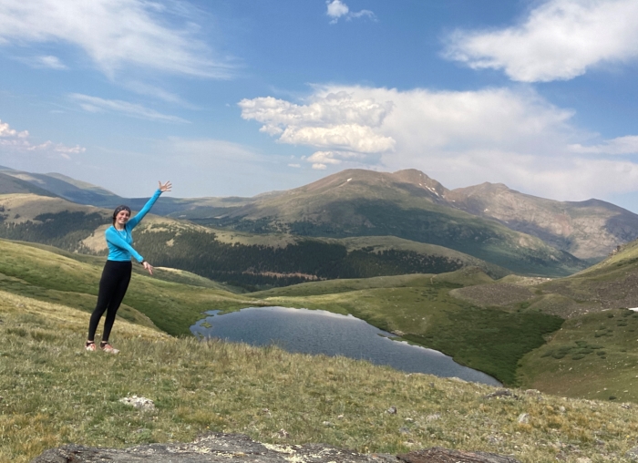 Photo of a beautiful mountain vista with Cameron posing in the near background, arms raised as if to show off the scenery.