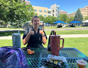 Ella sits at an outdoor picnic table across from the person taking the photo. She flashes two thumbs up.