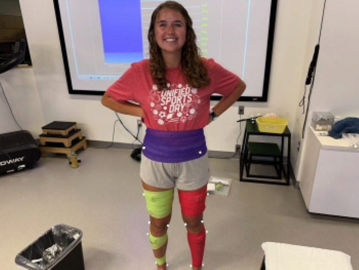 Photo of Ella standing and smiling. She is wearing wrapping around her waist and legs that will measure and analyze her gait movement.