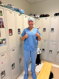 Ella, dressed in light-blue scrubs, flashes two thumbs up, ready to go into the OR