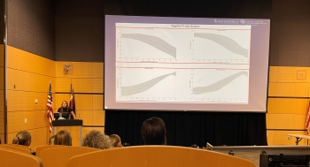 View from the back of an auditorium-style classroom with stadium seating and a huge projector screen at the front that is queued to a slide on Ella's presentation. Ella is standing on the left-hand side of the room behind a podium and is speaking to the audience.