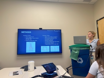 The photo shows the inside of a conference room with a TV mounted on the wall. There is a makeshift podium that used to simply be a recycling container but that now holds Josalin's laptop as she practices her presentation. Another intern looks on from the left of the photo.