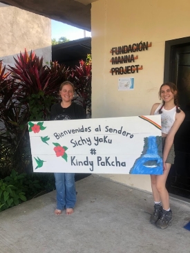 Emma and fellow intern Alexi stand facing the camera while holding up the sign they created for Javier's Sendero. It says "Bienvenidos al Sendero, Ichy yoKu, #, Kindy PaKcha"