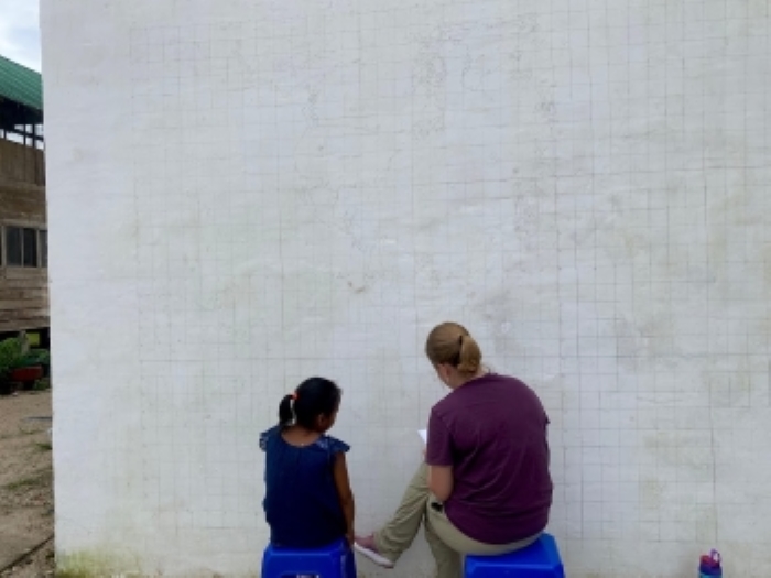 Emma and a young child sit side-by-side facing away from the camera, working on the drawing of a mural in Shandia, Ecuador.