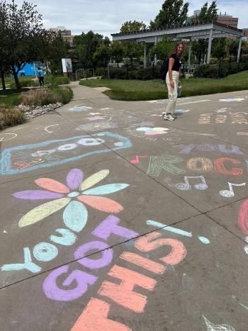 Lots of colorful pastel chalk art decorates a vast sidewalk courtyard area, and in the far-right background, Ella McLaughlin '26 can be seen standing.