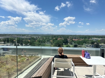 View of Ella from the back. She is sitting in a chair on a rooftop looking out at the vast cityscape view, with mountains barely visible in the horizon, partially obscured by clouds.