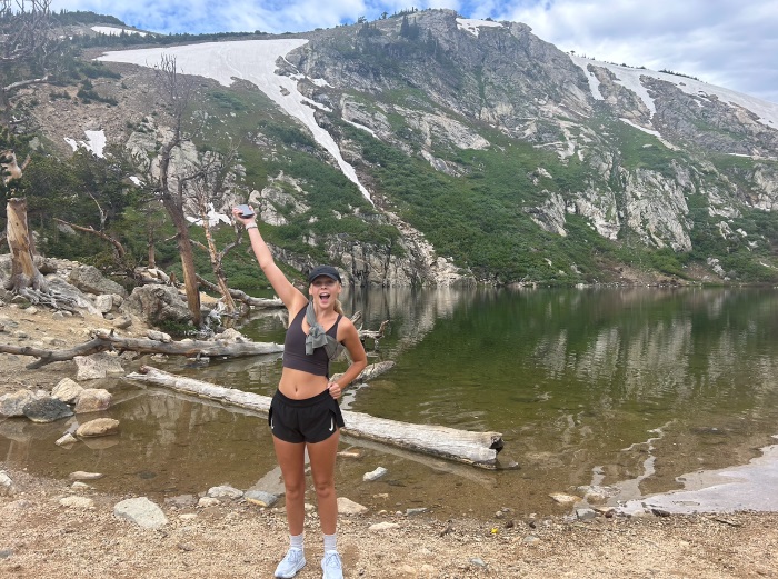 Josalin with a mountainous background. Her right arm is up as if to display the beautiful scenery. She is wearing black shorts and a crop top.