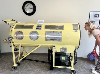 An old iron lung takes up most of the frame, with Josalin bent over at the waist on the right side of the iron lung and peering into it.