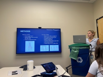 Josalin practicing a presentation behind blue recycling bin DIY podium in a small conference room