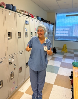 Josalin in a locker room wearing scrubs, including a plastic cap over her hair. She is flashing two thumbs up and smiling.