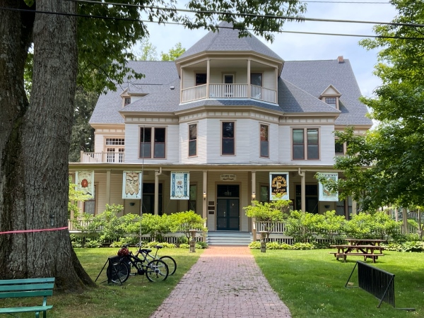 Large, three-story white house with expansive green lawn that has some wooden picnic tables set up in various spots. This is Alumni Hall at the Chautauqua Institute.