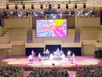 View of the stage and the Beach Boys performing. On the screen behind the musicians, "The Beach Boys" can be clearly seen in colorful font