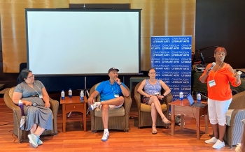 Photo of three people seated in armchairs on a stage and one person standing in front of an armchair with a microphone speaking