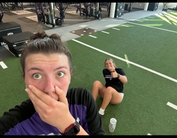 Krystin sits on the floor of a gym flashing a thumbs up sign while another person fakes surprise and takes this gym selfie.