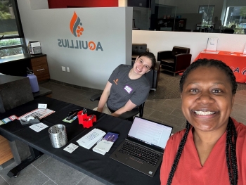 Kiana and another intern (who is taking the selfie) smiling from a desk space, the Aquillius logo visible but backwards in the background