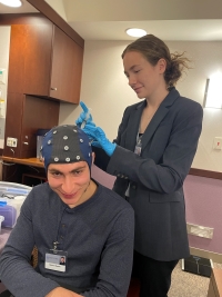 Laura stands over Moodi, applying a conductive goo while he wears an EEG cap with electrodes so she will be able to monitor his brain waves.