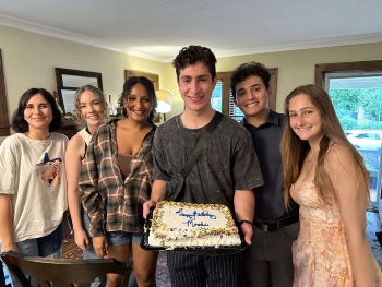 A group of six people huddles together with the third person from the right, Moodi Elawady, holding a birthday cake that says "Happy Birthday, Moodi!" in icing. Everyone is smiling.