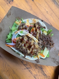 A cardboard boat contains two Korean tacos that appear to have shredded beef, lettuce, red onion, and a sauce drizzled over top.