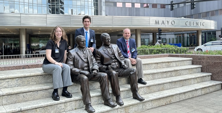 Moodi, Laura, and Dr. St. Louis sitting together on the steps with the Mayo Brothers statues in front of Mayo Clinic