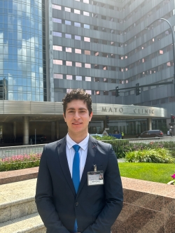 Moodi, dressed formally in a suit and tie, stands in front of the Mayo Clinic building, smiling.