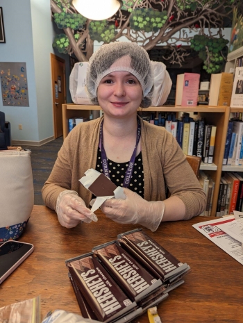 Nellie sits at a desk wearing plastic gloves and a plastic shower cap around her hair. Hershey's chocolate bars sit on the desk in front of her, and one is unwrapped.