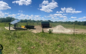 Photo of a sparse, grassy area with what looks like a solar panel and a water trough plus an unknown structure with a pile of giant sand to the right