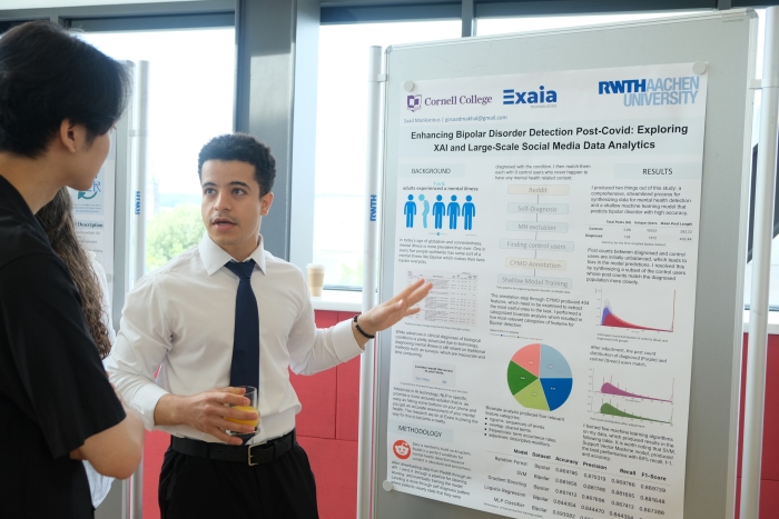 Saad, wearing a white collared shirt, tie, and dress pants, is in the middle of presenting his research poster to a curious onlooker who is facing toward Saad and away from the camera.