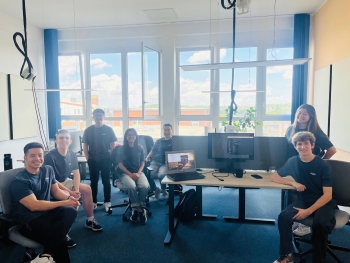 7 undergraduate students with matching black T-shirts that have small white text reading "RWTH" over the left breast are seated in a small computer lounge area, many seated in swivel chairs.