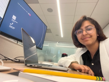 Smriti smiles from her desk, the angle of the photo looking up at her slightly. Her desk has a computer monitor and laptop visible on it.