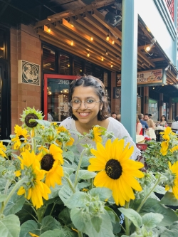 Smriti crouches, smiling, over a patch of sunflowers growing near a storefront.