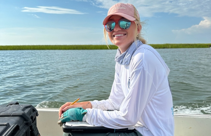 Anna, wearing a light-blue long-sleeved shirt and pinkish red hat, smiles at the camera. She is sitting in a boat.