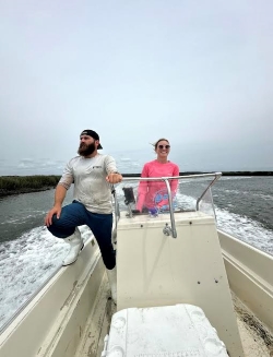 Anna driving a boat, a dark-beared man in a white tee and shorts to her right monitoring