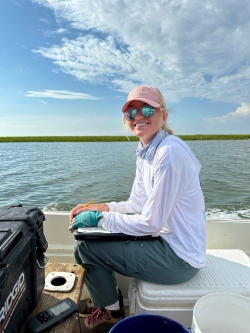 Anna, wearing a light-blue long-sleeved shirt and pinkish red hat, smiles at the camera. She is sitting in a boat.