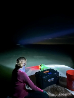 Anna driving a boat at night, bright rainbow colors near her steering area