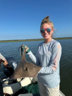 Anna smiles for the camera, holding up a stingray. She is wearing sunglasses and a light-blue top.