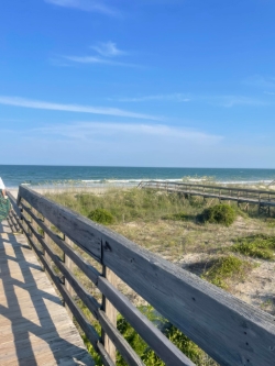 A wooden boardwalk with wooden railing goes out across a beachy area with some shrub grass