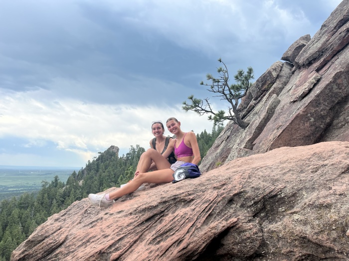 Alyza and her friend Ava sit on a flat plateaued rock that appears to be on the side of a mountain.