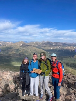 Alyza, Sophie, Zoe, and Milar at the summit of Mt. Bierstadt. They are holding a cardboard square that names the mountain and its altitude.