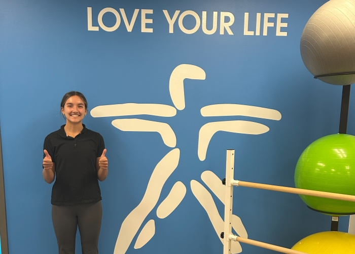 Alyza posing with a wall stenciling of a person extending their arms and legs in a jump with the words Love Your Life also in stenciling.