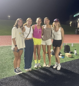 Group of five women with arms around each other in a group picture, seemingly on some sort of athletic field at night