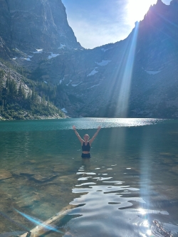 Alyza from afar, waist-high in mountain lake water, her arms extended over her head in a gesture of excitement and vitality
