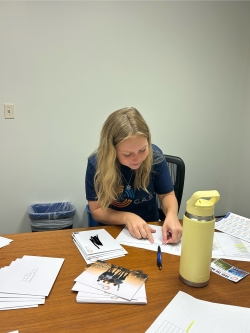 Brynn works to fold a thank-you card. She is seated at a brown table strewn with papers and folded cards and other writing and cutting materials. She wears a blue T-shirt with the letters C.A.S.T. printed prominently on the chest.