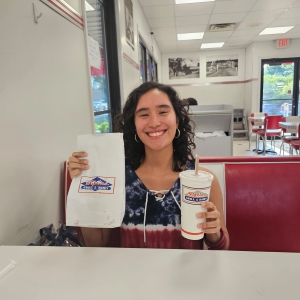 Photo of Claudia in a diner booth holding up her drink in what looks like a styrofoam cup and the paper bag of food as well